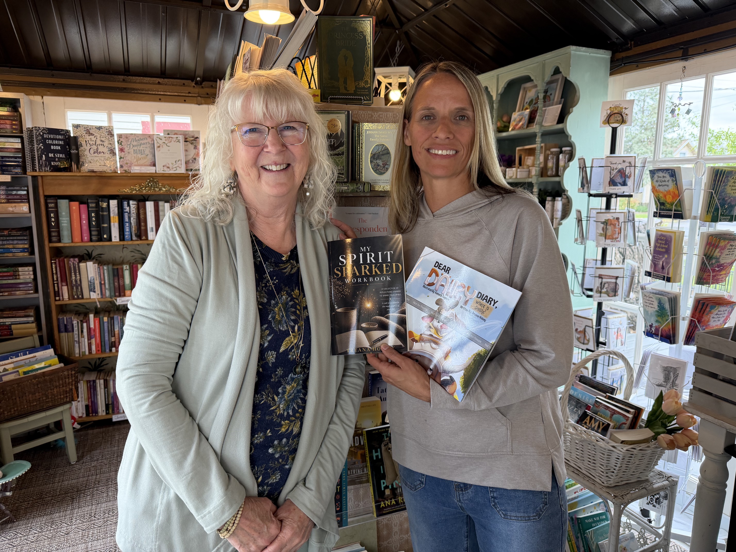 Two smiling visitors holding books among the shelves, cards, and gifts at House of the Book