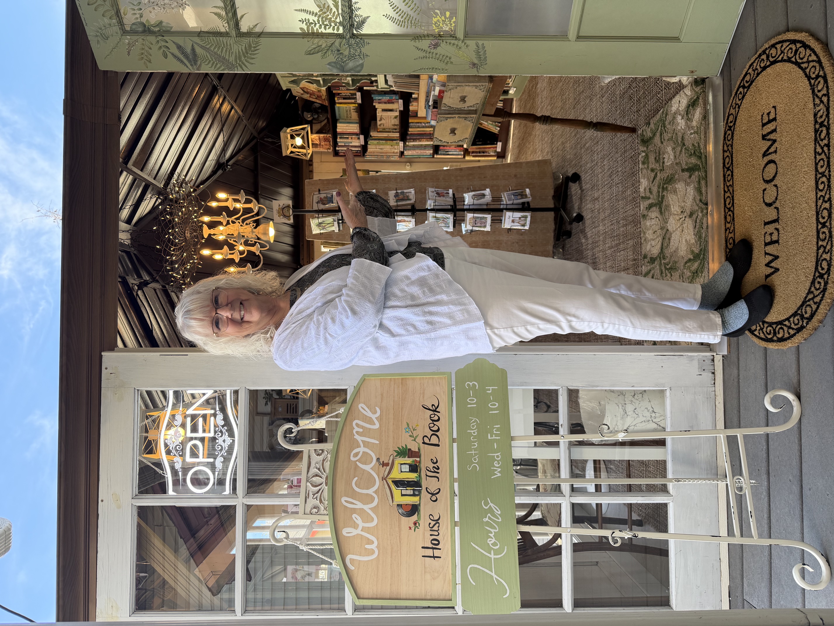 A bookseller welcomes visitors at the open doorway of House of the Book, beside a welcome sign and a view into the shop